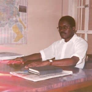 José Carlos do Nascimento, ainda jovem e usando camisa social, sentado à uma mesa de escritório cercado de livros e cadernos, em um momento de profunda concentração e estudo.